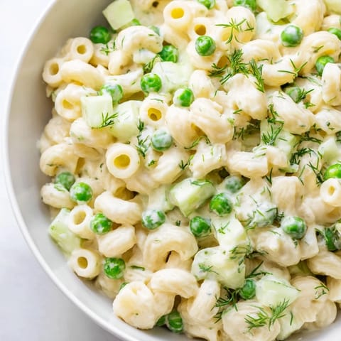 A close-up view of Creamy Dill Pickle Pasta Salad in a white bowl, showcasing ditalini pasta, diced pickles, and sweet peas coated in a rich, tangy dressing, garnished with fresh dill.